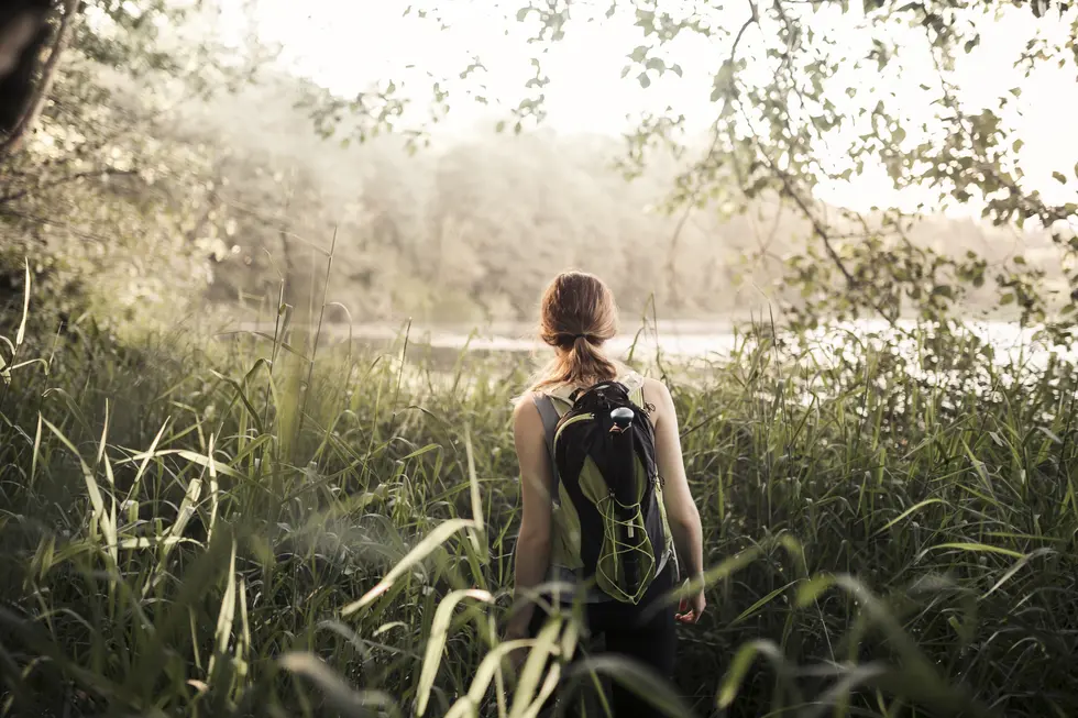 Frau mit Rucksack, die durch hohes Gras in einer naturnahen Umgebung mit Bäumen und einem Gewässer im Hintergrund wandert, in sanftem Licht.