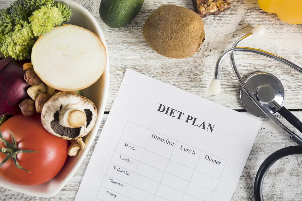 Bunte Auswahl an frischem Gemüse und Obst in einer Schüssel zusammen mit einem Diet-Plan auf einem Holztablett, das die Tabelle für Mahlzeiten der Woche zeigt, ergänzt durch ein Stethoskop, das auf das Dokument gelegt ist, um Gesundheit und Ernährung zu symbolisieren.