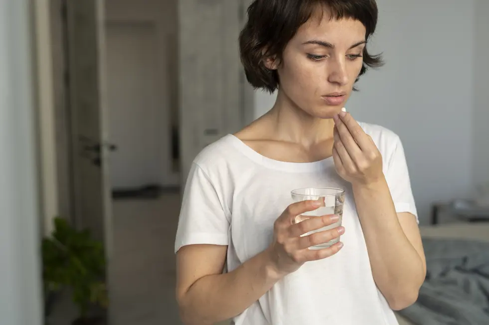 Eine besorgte Frau steht in einem modernen Raum und hält ein Glas Wasser in der linken Hand, während sie mit der rechten Hand eine Pille zwischen Daumen und Zeigefinger aufnimmt. Ihr nachdenklicher Gesichtsausdruck deutet darauf hin, dass sie sich auf das Einnehmen des Medikaments vorbereitet. Der Raum ist hell und freundlich gestaltet, mit einer Pflanze im Hintergrund, was eine entspannte Atmosphäre schafft.