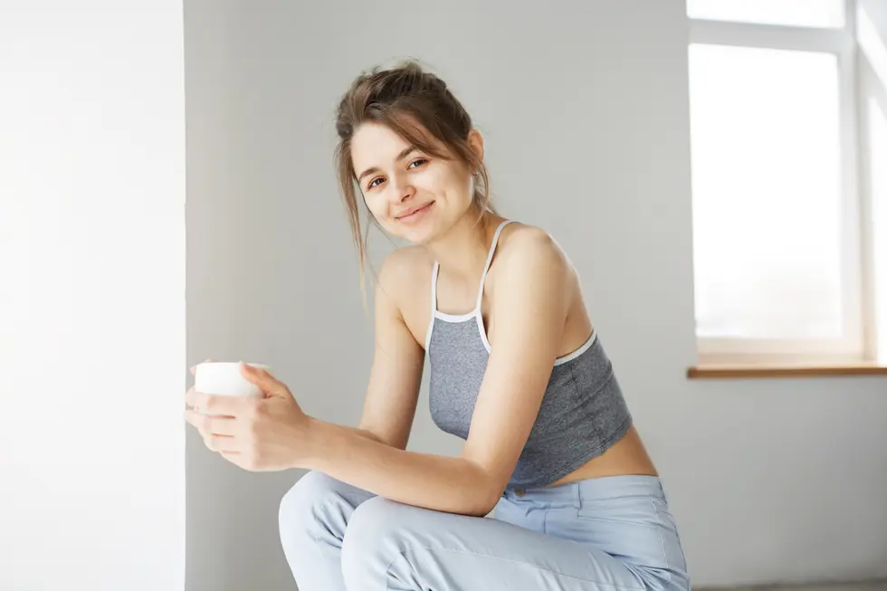 Junge Frau mit einem Glas in der Hand, die an einer Wand sitzt und lächelt. Sie trägt ein sportliches Top und bequeme Hose, während Tageslicht durch das Fenster auf sie fällt. Der Hintergrund ist hell und neutral gehalten, was eine entspannte Atmosphäre schafft.