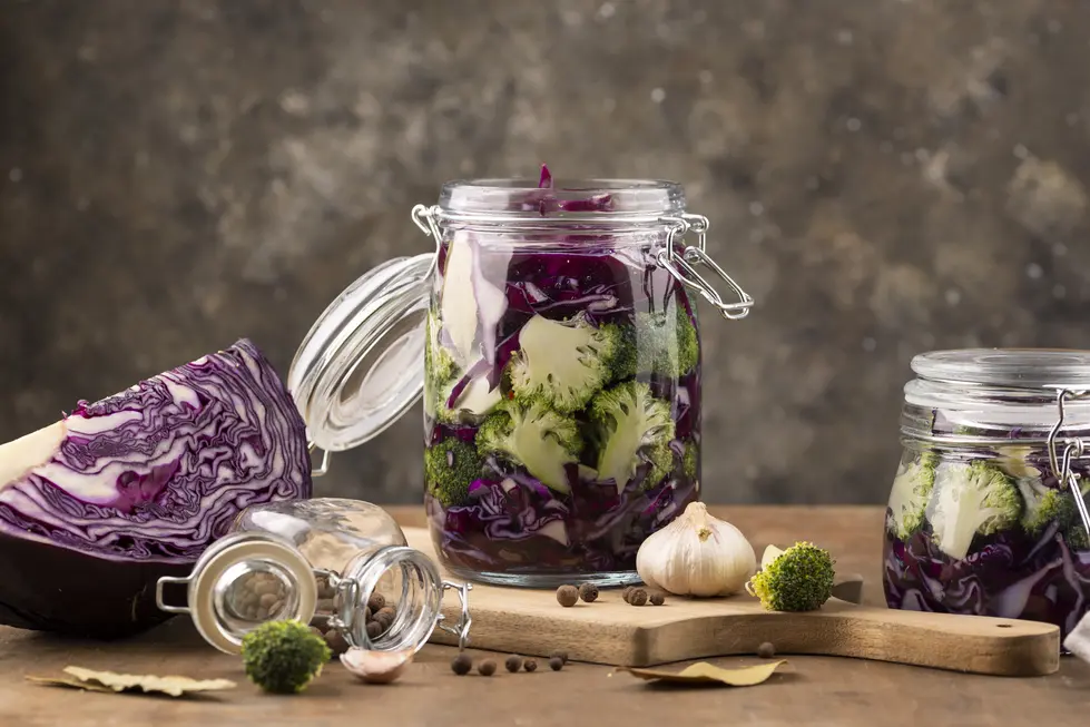 Glasgefäße mit fermentiertem Gemüse, darunter lila Kraut und Brokkoli, auf einem Holzbrett arrangiert. Neben den Gläsern liegen frischer Knoblauch und Gewürze wie Pfefferkörner. Das Bild zeigt die gesundheitsfördernde Zubereitung von fermentierten Lebensmitteln, die für die Unterstützung der Darmgesundheit und die Stärkung des Mikrobioms bekannt sind.