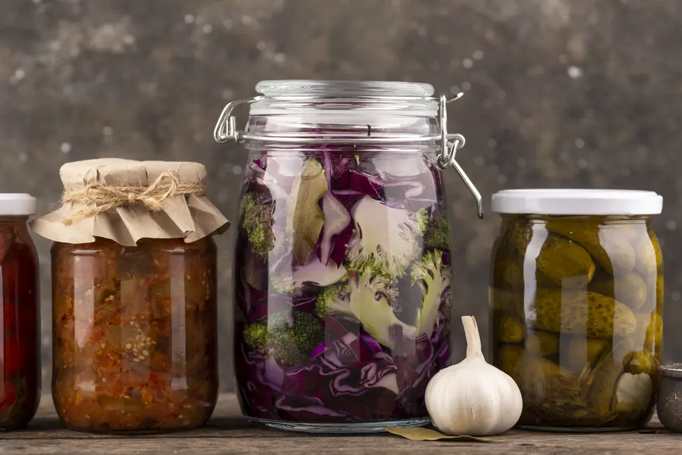 Fermentiertes Gemüse, darunter rote Kraut mit Brokkoli und Gewürzen in einem Glasbehälter, ziert einen rustikalen Holztisch. Neben dem Glas befinden sich ein weiteres eingekochtes Gemüse in einem Glas mit Papierabdeckung und ein Glas mit eingelegten Gurken. Eine Knoblauchzehe liegt ebenfalls auf dem Tisch, was das Bild zu einer ansprechenden Darstellung gesunder, hausgemachter Lebensmittel macht.