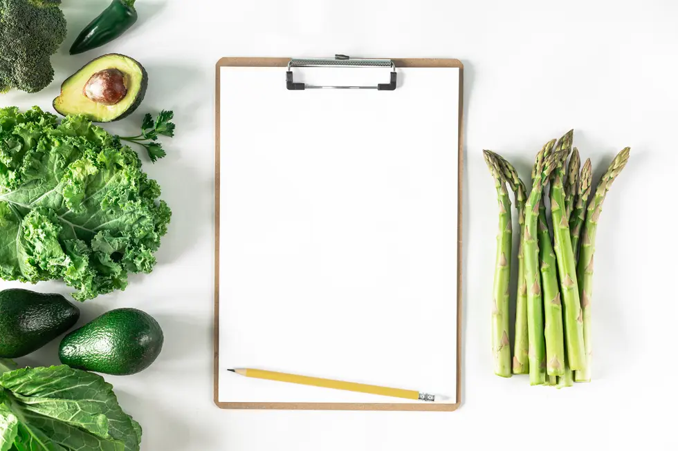 Eine Auswahl an frischem Gemüse, darunter Brokkoli, Avocado, grünes Blattgemüse und Spargel, liegt zusammen mit einem leeren Notizblock und einem gelben Bleistift auf einem weißen Tisch.