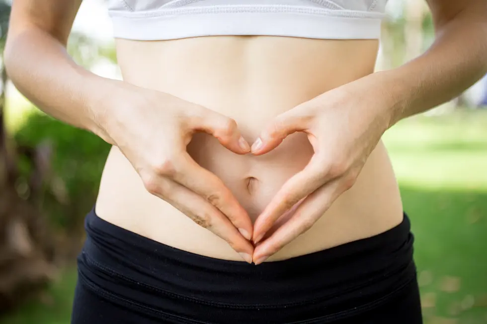 Frau formt mit ihren Händen ein Herz vor ihrem Bauch, trägt einen Sport-BH und Leggings, steht im Freien mit grünem Hintergrund, symbolisiert Gesundheit und Selbstpflege in Bezug auf die Darm-Haut-Achse.