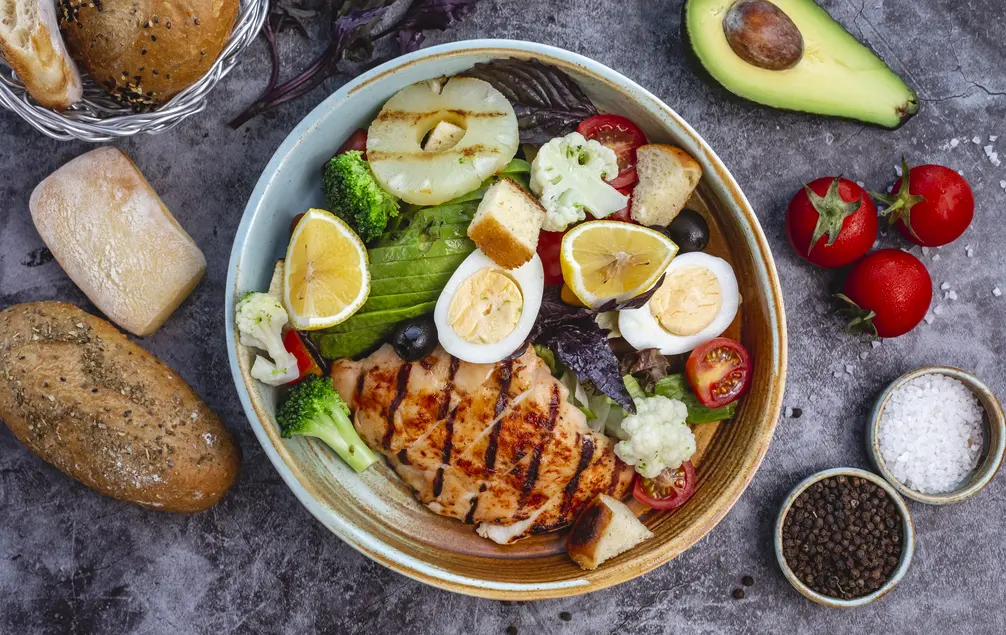 Gesunde Mahlzeit mit gegrilltem Hähnchen, buntem Gemüse, Hartgekochten Eiern, Zitronenscheiben und frischen Kräutern. Dazu Brote in verschiedenen Sorten und Gewürze wie Salz und Pfeffer. Ideal für eine ausgewogene Ernährung.