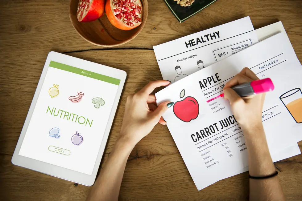 Eine Person notiert Informationen über Ernährung auf einem Informationsblatt, während ein Tablet mit einem grünen Bildschirm und verschiedenen Lebensmittelsymbolen neben ihr liegt. Auf dem Tisch befinden sich frisches Obst in einer Schale sowie mehrseitige Dokumente mit Nährwertangaben zu Äpfeln und Karottensaft. Der Fokus liegt auf der gesunden Ernährung und der Analyse von Nährstoffen.