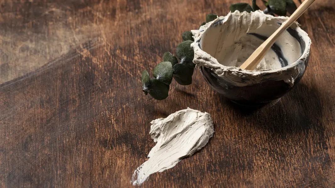 Schüssel mit Heilerde in einem natürlichen Farbton auf einem Holztisch, mit einem Holzspatel. Neben der Schüssel liegt ein Löffel Heilerde, und eine grüne Pflanze ist sichtbar, die dem Bild eine natürliche Note verleiht.