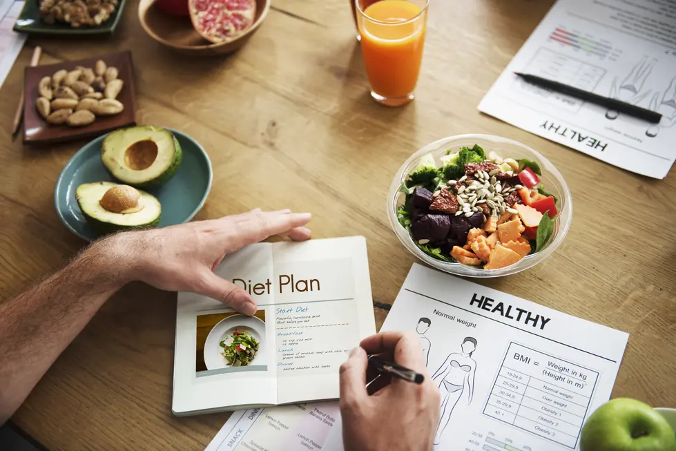 Person plant eine gesunde Ernährung, indem er einen Diätplan aus einem Heft studiert und Notizen macht. Auf dem Tisch sind frische Zutaten zu sehen, darunter Avocado, ein bunter Salat mit verschiedenen Gemüsesorten, sowie Snacks wie Nüsse und ein Glas frisch gepresster Saft. Rechts sind Gesundheitsunterlagen sichtbar, die Informationen zur Gewichtskontrolle und BMI-Berechnung enthalten.