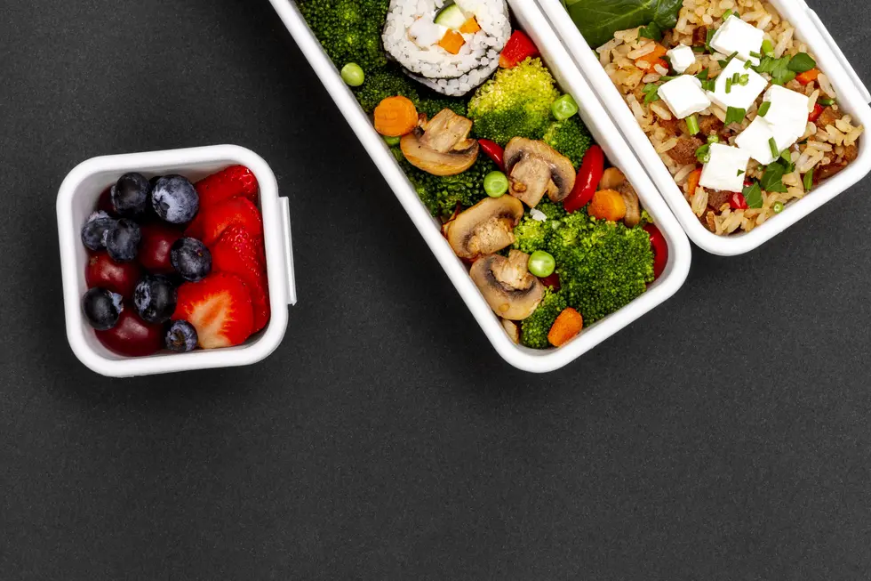 Bunte Meal-Prep-Boxen mit gesundem Essen: Eine Box enthält frische Erdbeeren, Blaubeeren und Trauben, während die anderen Boxen gedämpftes Gemüse wie Brokkoli, Karotten und Champignons sowie eine Portion Reis mit Feta und frischen Kräutern zeigen. Das Arrangement befindet sich auf einer dunklen Oberfläche.