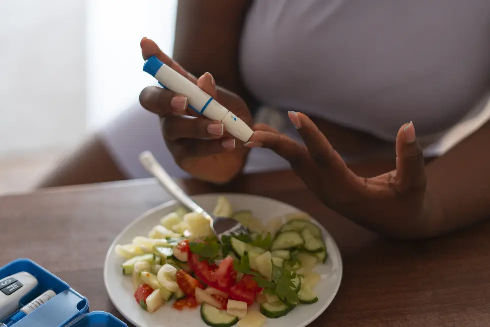 Frau misst ihren Blutzuckerspiegel mit einem Blutmessgerät, während sie eine gesunde Gemüsesalat-Mahlzeit mit Gurken, Tomaten und frischen Kräutern vor sich hat. Auf dem Tisch liegt ein medizinisches Utensil zur Blutzuckermessung.