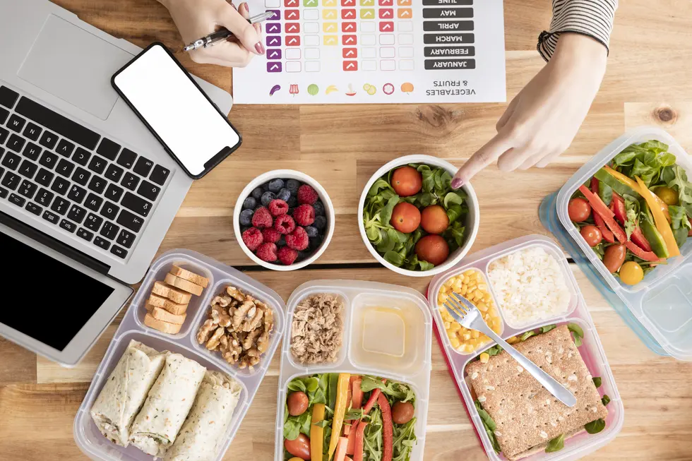 Gesunde Meal-Prep-Boxen mit proteinreichen Zutaten und frischem Gemüse, darunter Rucola, Tomaten, Blaubeeren und Himbeeren. Auf dem Tisch befindet sich ein Laptop, ein Smartphone sowie ein Blatt mit einem Essensplan, das die Planung der Ernährung unterstützt.