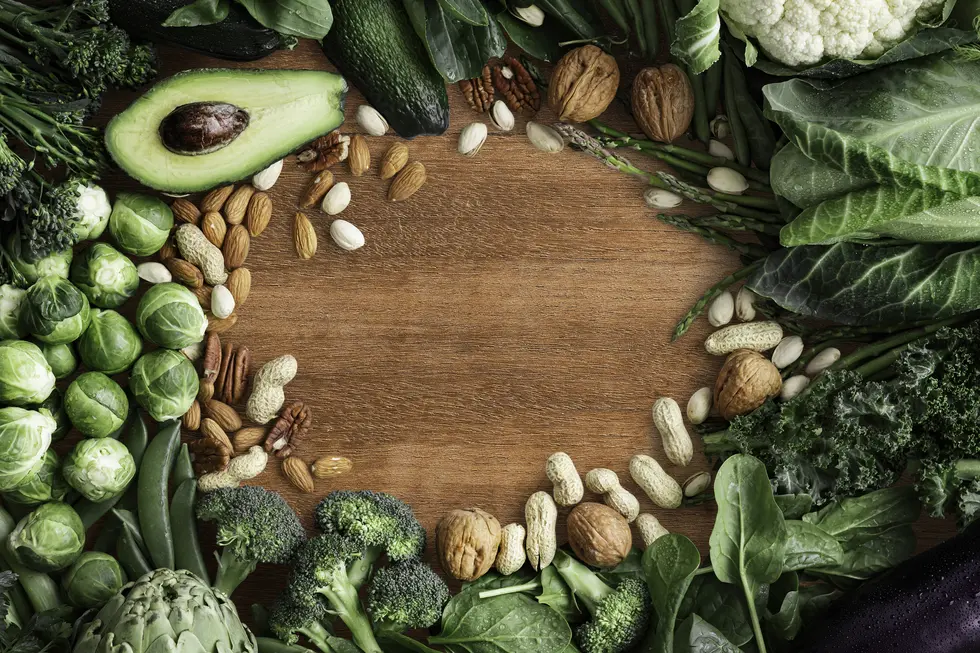 Verschiedene grüne Gemüsesorten und Nüsse, einschließlich Avocado, Brokkoli, Pekannüsse, Mandeln und Rosenkohl, sind auf einem Holzbrett dekorativ angeordnet, um eine gesunde Ernährung zu betonen.