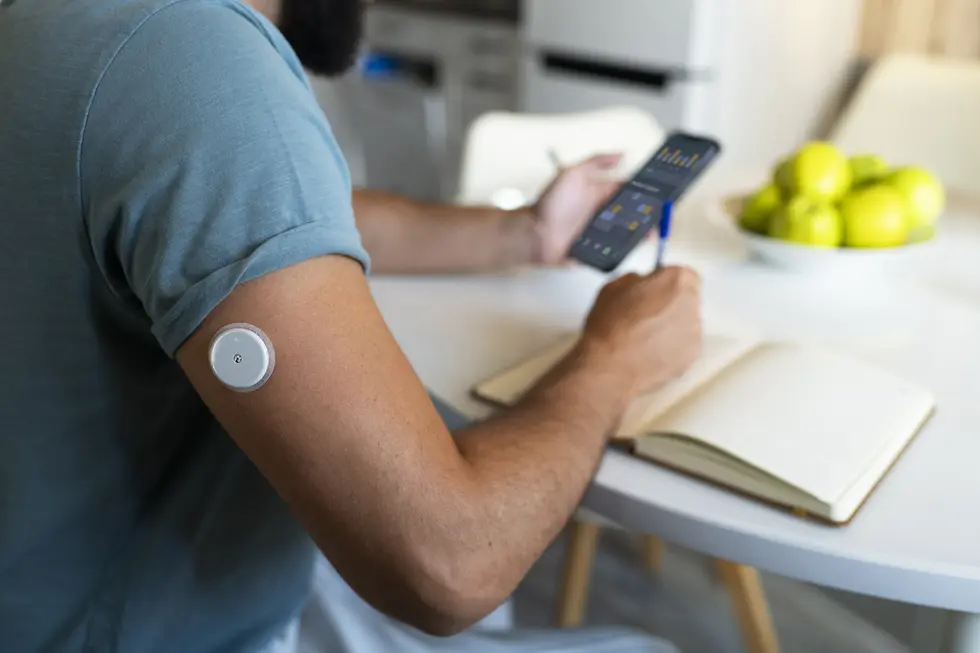Mann mit Glukosemonitor auf dem Oberarm, der am Tisch sitzt, ein Smartphone in der Hand hält und Notizen in ein Notizbuch macht. Im Hintergrund sind grüne Äpfel in einer Schale sichtbar.