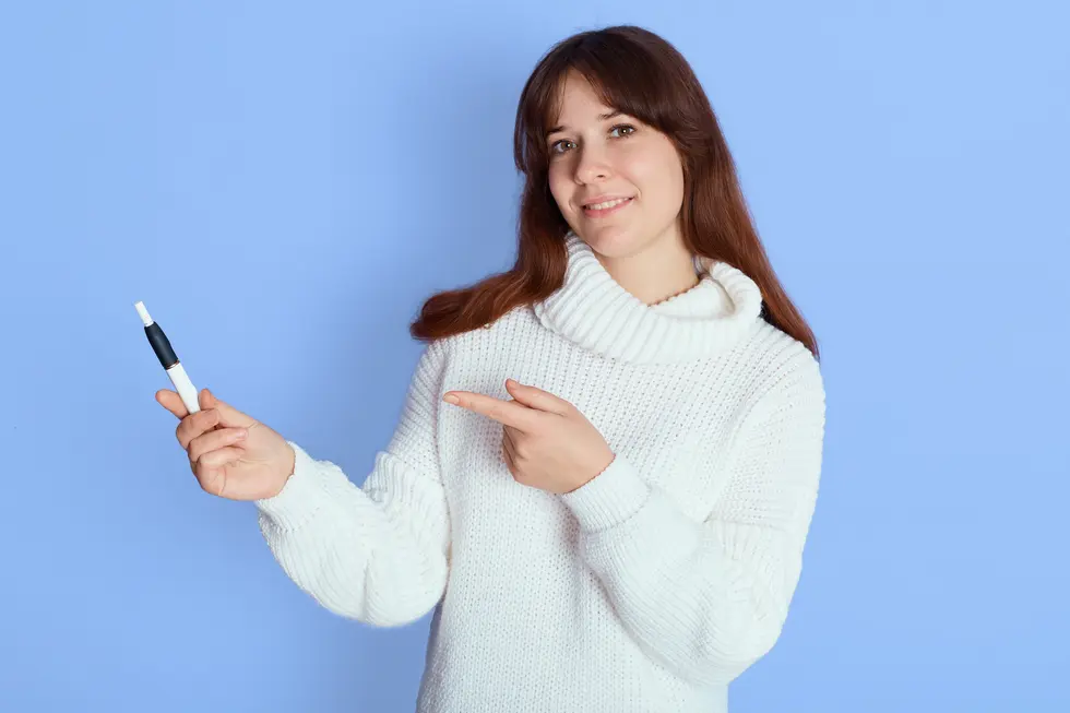 Frau in einem weißen Pullover, die mit einem Lächeln in die Kamera schaut und mit einem Finger auf einen Stift zeigt, vor einem hellblauen Hintergrund.