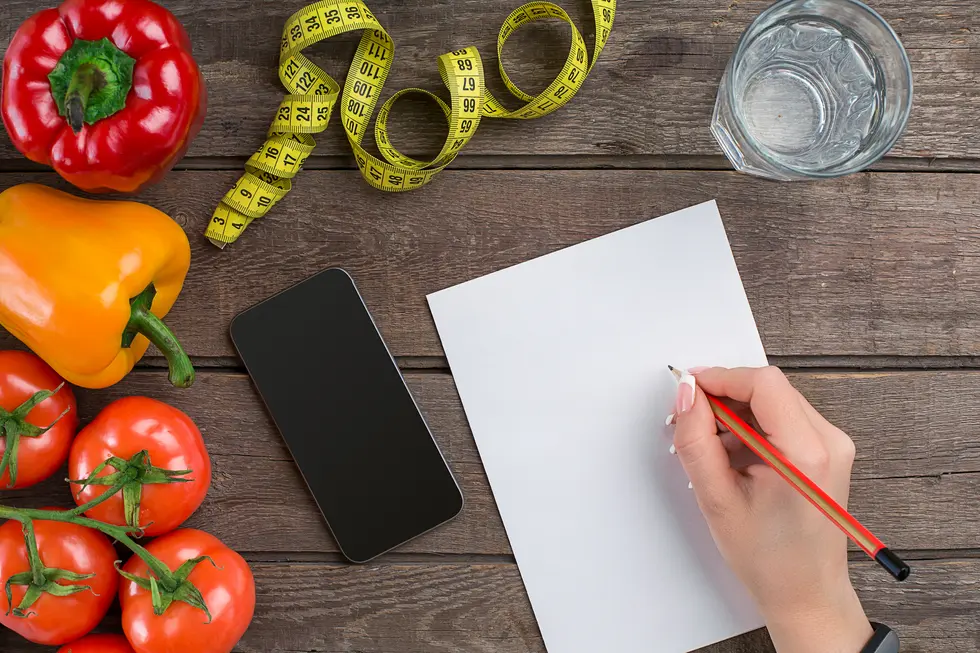 Gesunde Lebensmittel wie Paprika und Tomaten liegen auf einem Holztisch neben einem Smartphone und einem Glas Wasser. Eine Hand hält einen Bleistift über ein leeres Blatt Papier, während ein Maßband daneben liegt, was auf eine aktive Planung oder dokumentierte Ernährung hinweist.