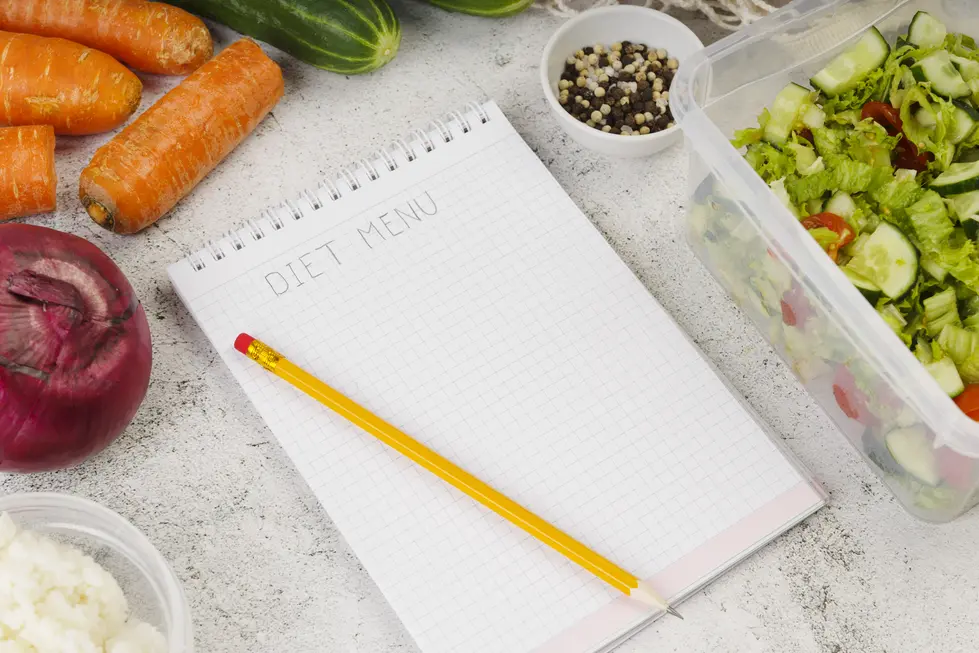Frisches Gemüse, darunter Karotten, Gurke und rote Zwiebel, liegen neben einem Notizblock mit der Aufschrift "DIET MENU" und einem gelben Bleistift. Eine Schüssel mit einem bunten Salat und Gewürzen ist ebenfalls sichtbar. Das Bild vermittelt den Eindruck von gesunder Ernährung und der Planung von Mahlzeiten.