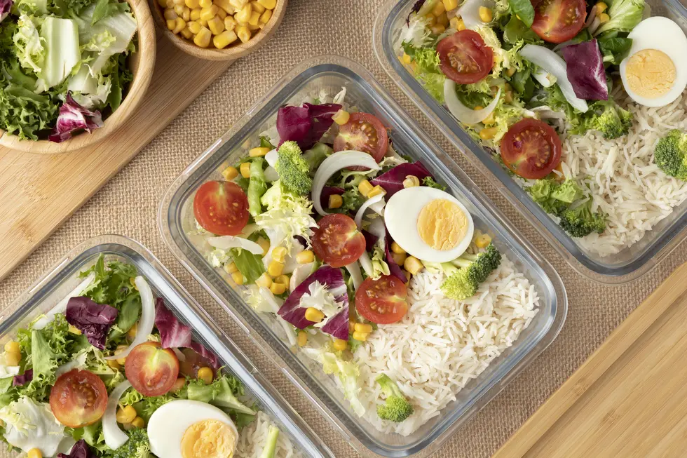 Vorbereitete Mahlzeiten in Glasbehältern mit einer bunten Mischung aus frischem Salat, Brokkoli, Kirschtomaten, Mais und gekochtem Ei, serviert mit Reis. Im Hintergrund eine Schüssel mit grünem Salat und eine weitere Schüssel mit Mais.