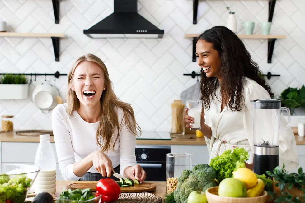 Zwei Frauen stehen in einer modernen Küche und haben sichtbar Spaß beim Kochen. Eine Frau, die ein weißes Langarmshirt trägt, schneidet frisches Gemüse auf einem Holzbrett, während sie lacht. Die andere Frau, gekleidet in einem leichten, hellen Oberteil, hält ein Glas mit einem Getränk in der Hand und blickt lächelnd auf ihre Freundin. Rund um sie befinden sich verschiedene gesunde Lebensmittel, darunter frisches Gemüse wie Brokkoli und Salat, sowie Obst in einer Schüssel. Die Küchenzeile ist hell und einladend gestaltet, mit offenen Regalen und einer neutralen Farbpalette, was eine harmonische Atmosphäre schafft. Dies verdeutlicht die Freude am gesunden Kochen und die Bedeutung einer ausgewogenen Ernährung.