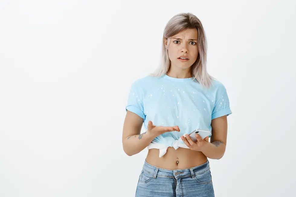 Junge Frau mit kurzem graublonden Haar, die in einem hellblauen T-Shirt und Jeans bekleidet ist, schaut überrascht oder verwirrt auf ihr Smartphone und hebt dabei eine Hand in Fragegestellung. Der Hintergrund ist neutral und hell, was ihre Mimik und Haltung hervorhebt.
