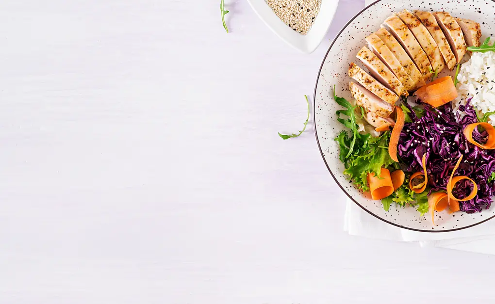 Gesunde Mahlzeit mit gegrilltem Hähnchenbrustfilet, serviert auf einem bunten Salatbett aus grünem Blattgemüse, violettem Kohl, und Karottenraspeln, ergänzt mit einer Portion Reis und Sesam. Ideal für eine ausgewogene Ernährung.