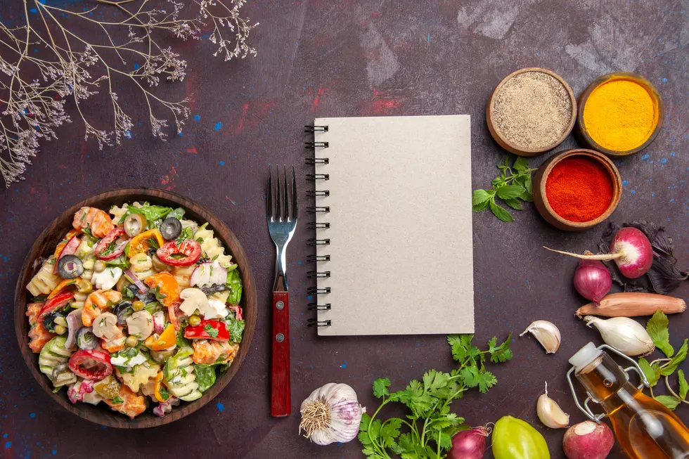 Bunte Salatschüssel mit verschiedenen Gemüsesorten, Kräutern, Essigflasche und Gewürzen auf einem Tisch, daneben ein leeres Notizbuch und eine Gabel. Ideal für Meal Prep und Rezeptplanung.