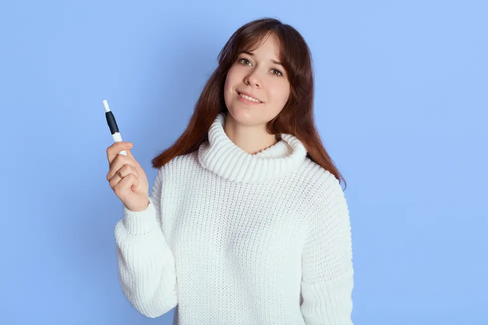 Frau in einem weißen Pullover mit Rollkragen, die mit einem Produkt in der Hand lächelt, vor einem blauen Hintergrund.