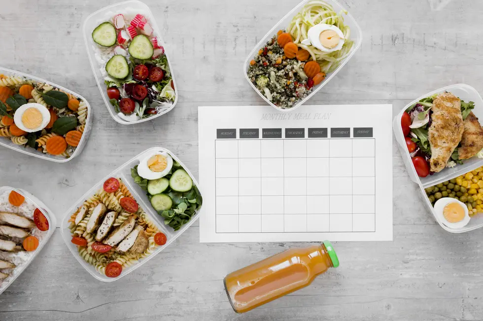 Gesunde Mahlzeiten in Aufbewahrungsboxen, die verschiedene bunte Gerichte enthalten, darunter Pasta, Salate und gekochte Eier, angeordnet um einen weißen monatlichen Essensplan. Ein orangefarbener Smoothie oder Saft befindet sich unten im Bild.