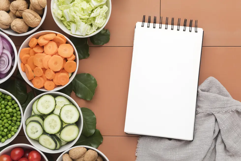Gesunde Zutaten in Schüsseln, darunter geschnittene Karotten, Gurken, rote Zwiebeln, Erbsen und Salat, umgeben ein leeres Notizbuch auf einem terrakottafarbenen Untergrund, das eine kreative Planung von Rezepten und Ernährungsplänen für eine insulinresistente Ernährung symbolisiert.