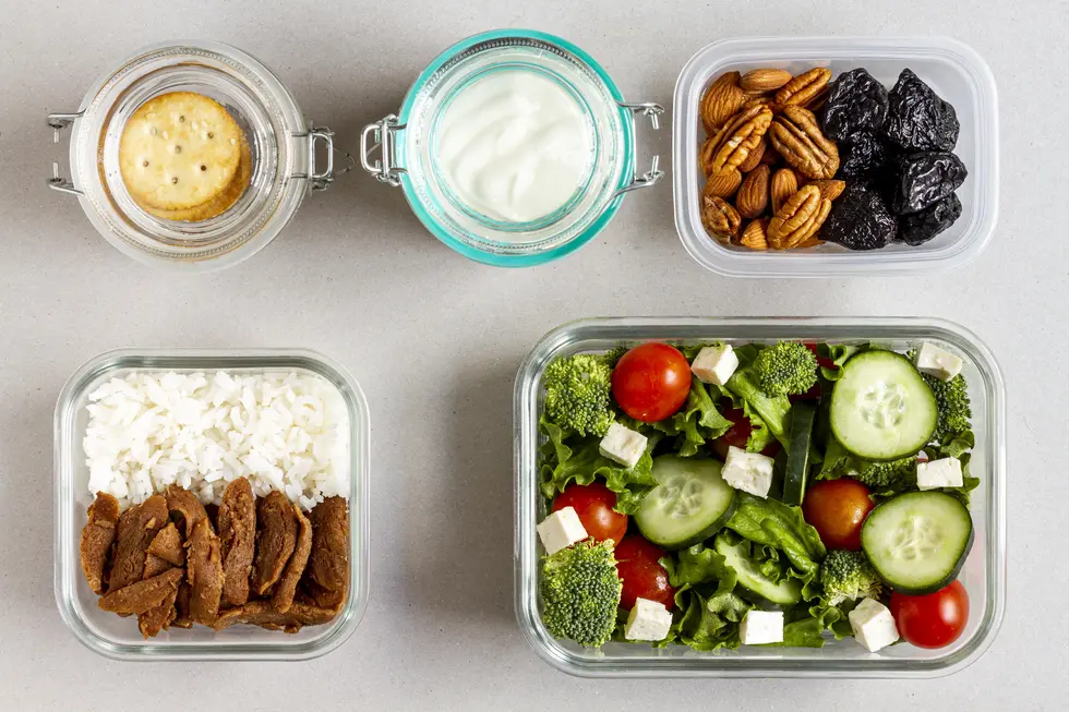 Glasbehälter mit abwechslungsreicher Mahlzeitenvorbereitung: Gemischter Salat mit Tomaten, Gurken, Brokkoli und Feta, dazu ein Portion Reis mit pflanzlichem Eiweiß, sowie Snacks in separaten Gläsern: ungesüßter Joghurt und Nüsse. Gesundes Meal Prep für eine ausgewogene Ernährung.