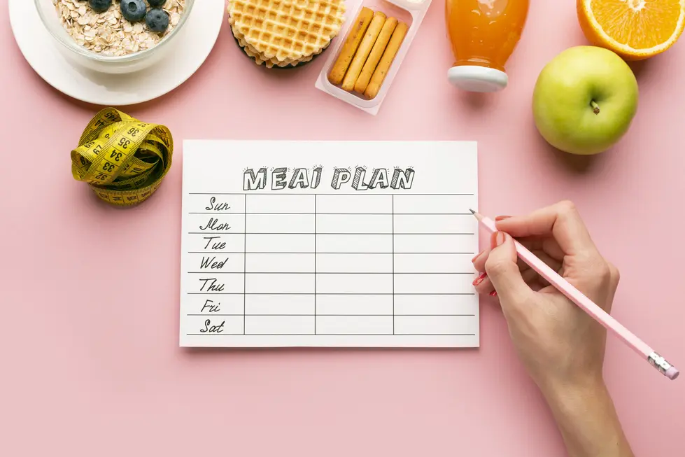 Eine Hand, die einen Zettel mit der Überschrift "MEAL PLAN" und verschiedenen Zeilen für die Wochentage hält, umgeben von gesunden Lebensmitteln wie einem Apfel, einer Orange, Haferflocken mit Beeren, Waffeln, Snacks und einem Getränk, auf einem rosa Hintergrund.
