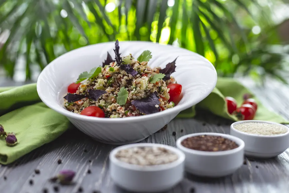 Bunte Quinoa-Bowl mit frischen Tomaten, Kräutern und Gemüse auf einem weißen Teller, umgeben von kleinen Schalen mit verschiedenen Körnern auf einem rustikalen Holzuntergrund.